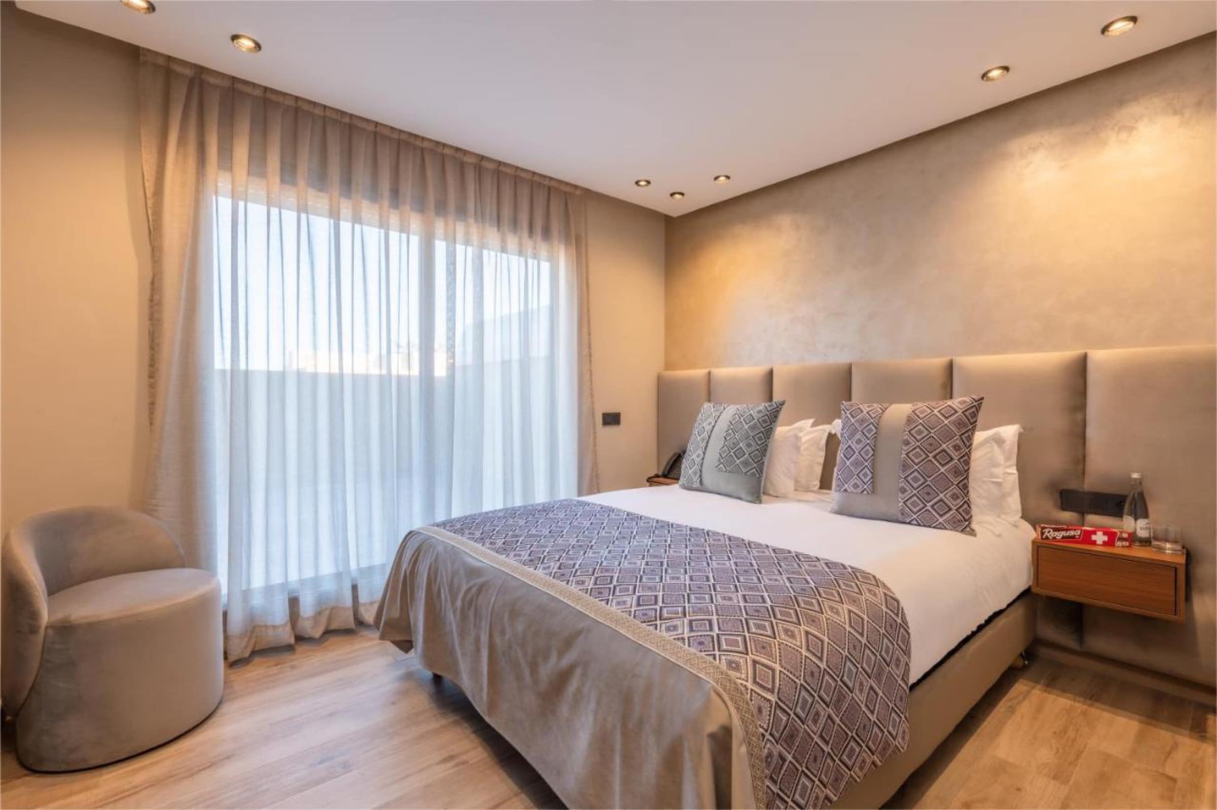 Une chambre taupe, tête de lit capitonnée et fenêtre voilée
